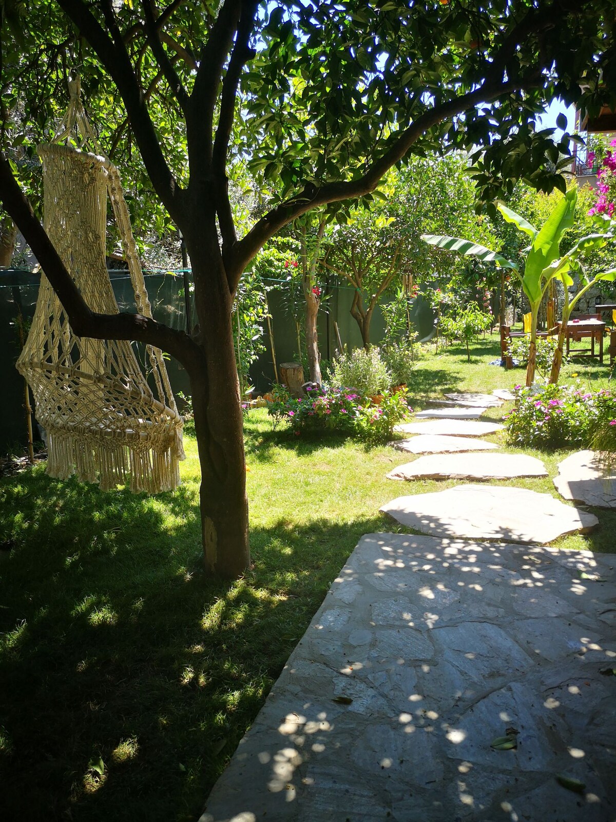 A serene garden pathway is lined with smooth stones, leading through lush greenery and colorful flowering plants. A macramé hammock hangs from a tree, inviting relaxation amid the natural surroundings. The vibrant garden is framed by trees, creating a peaceful retreat.