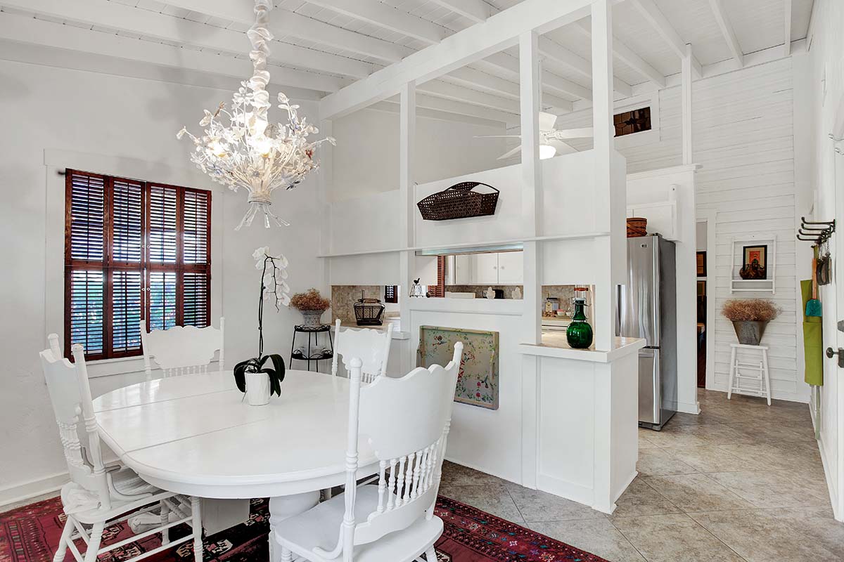 A bright dining area features a large round white table surrounded by multiple white chairs. A unique chandelier adds a decorative touch above the table. Light filters through wooden shutters, and the space is separated from the kitchen by a small partition, enhancing an open layout.