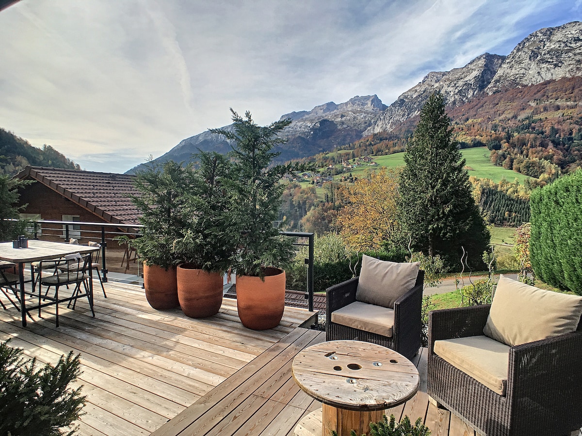 A spacious terrace features a wooden deck, complemented by comfortable seating and large potted trees. Scenic views of rolling hills and mountains are visible in the background, showcasing the natural landscape surrounding the chalet.