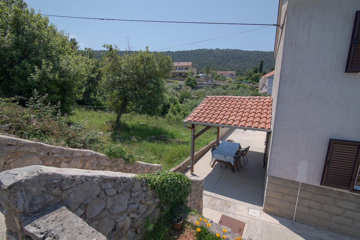 A view of the exterior reveals a spacious terrace with a tiled roof and a dining table set for outdoor meals. Lush greenery and trees fill the surrounding area, with a gently sloping landscape leading to distant hills.