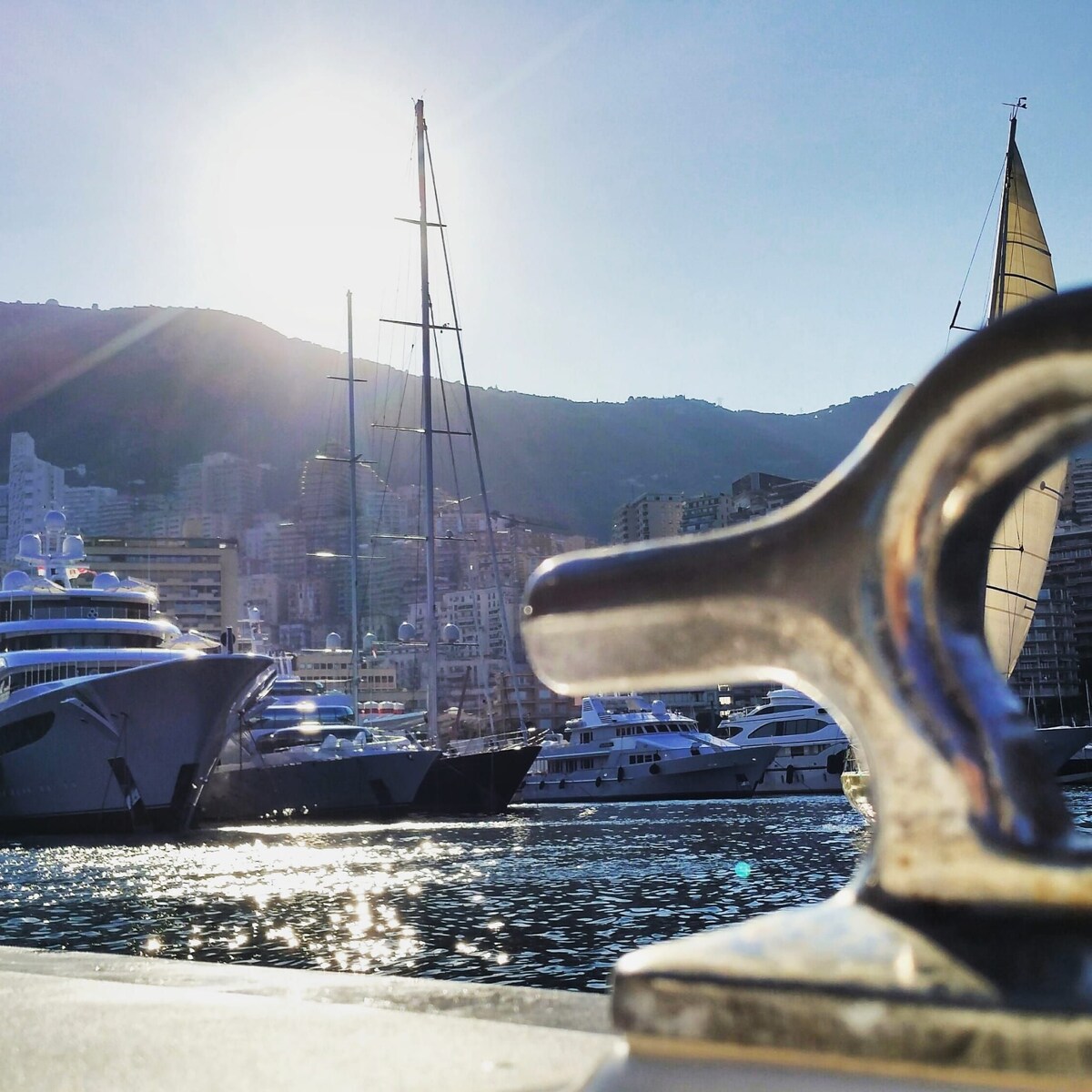The image showcases a view of boats docked in the harbor, with a glistening water surface reflecting sunlight. A picturesque backdrop of the mountains and city buildings complements the scene. The foreground features a metal cleat, emphasizing the maritime setting.
