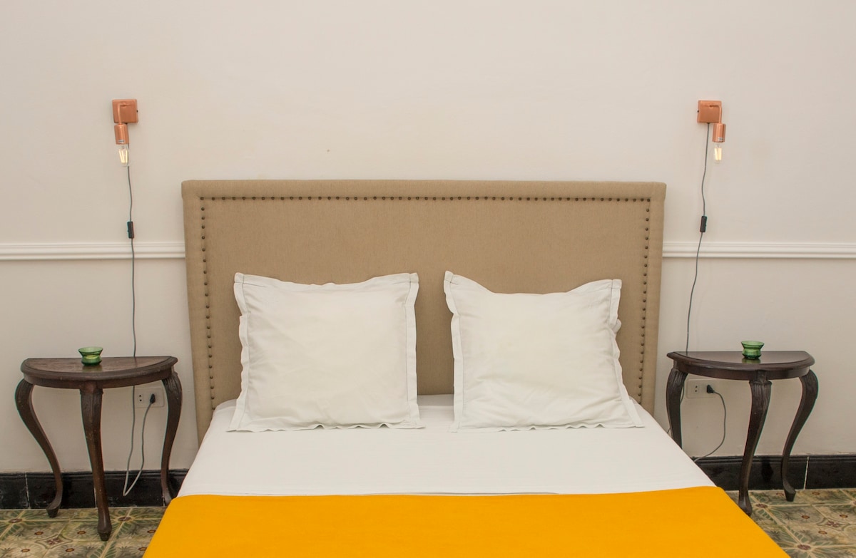 A spacious bed is positioned against a neutral wall, complemented by two decorative nightstands. Each nightstand features a small candleholder. Soft white pillows create a clean and inviting look, while a vibrant yellow throw adds a touch of color.