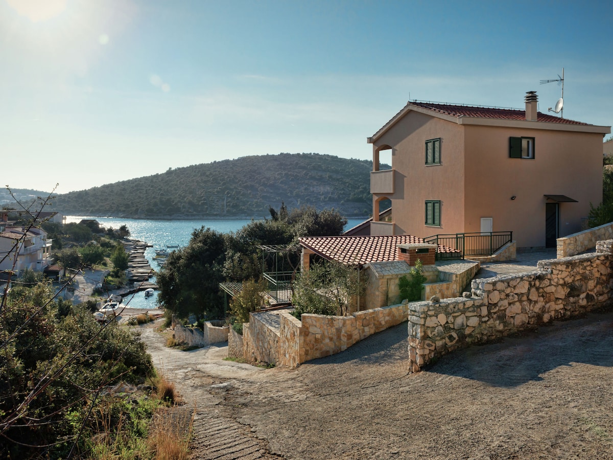 A three-story building is situated on a sloping hillside, offering a scenic view of the bay. The exterior features soft peach-colored walls and a terracotta roof, surrounded by lush greenery and stone pathways. Calm waters glisten in the distance under a clear blue sky.