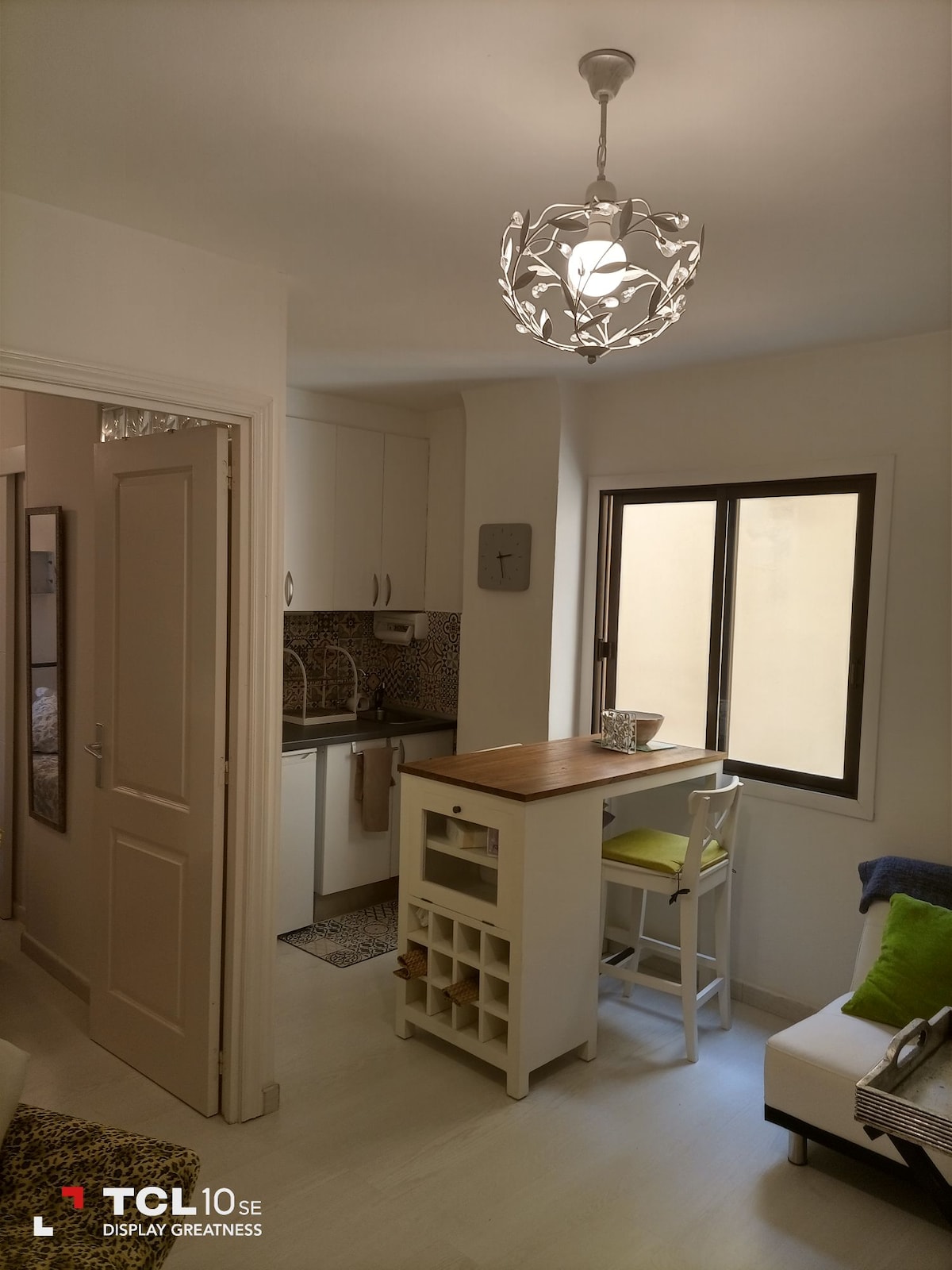 A compact kitchen area is visible, featuring light-colored cabinetry and patterned backsplash. A wooden countertop serves as a casual dining space, accompanied by a tall white chair. Natural light enters through a window, illuminating the room's neutral tones and accented with decorative green cushions.