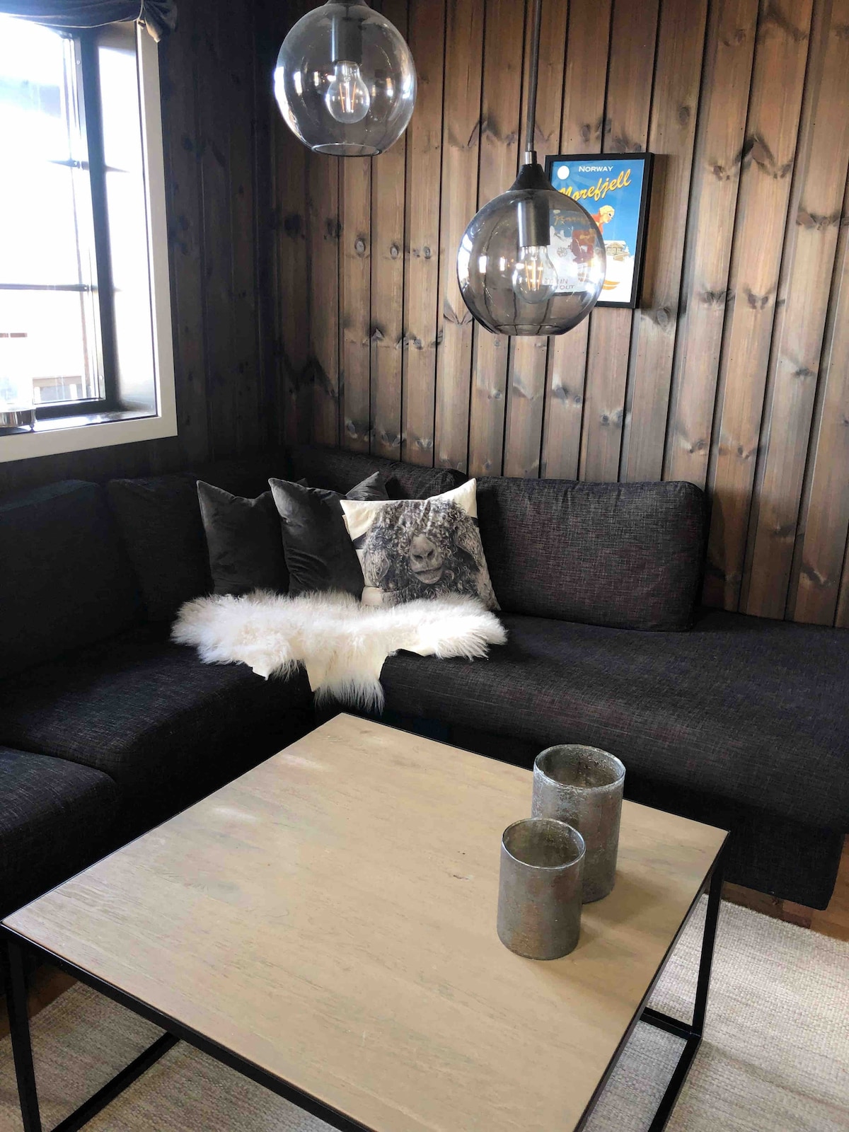 A cozy seating area features a dark sectional couch with accent pillows, including one displaying a graphic design. A soft throw is positioned casually on the couch. A light wood coffee table sits in front, complemented by two metallic vases. Natural light streams through the window.