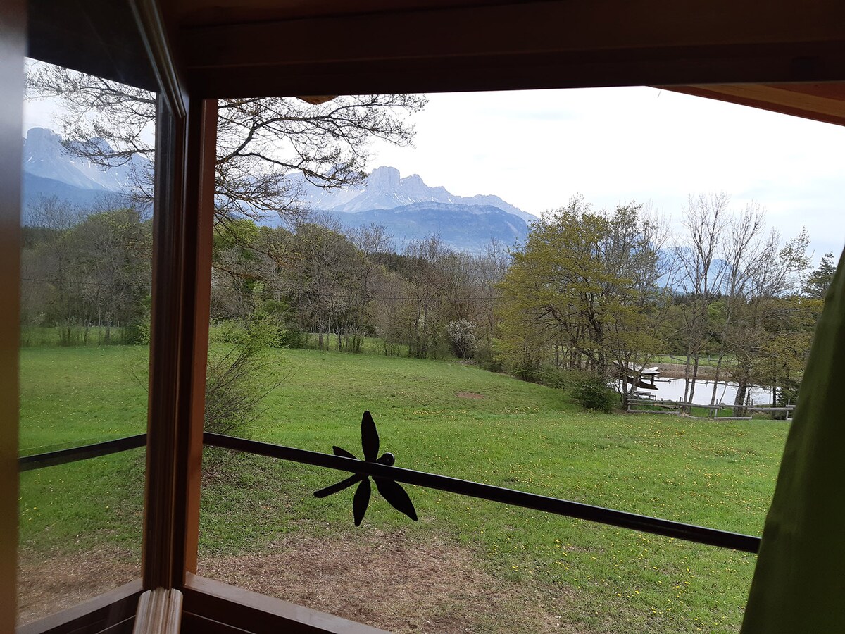 The view from a window offers a serene landscape featuring a grassy field, trees, and distant mountains. A small pond is visible in the background, enhancing the natural setting. The window frame includes decorative elements, adding a touch of charm to the scene.