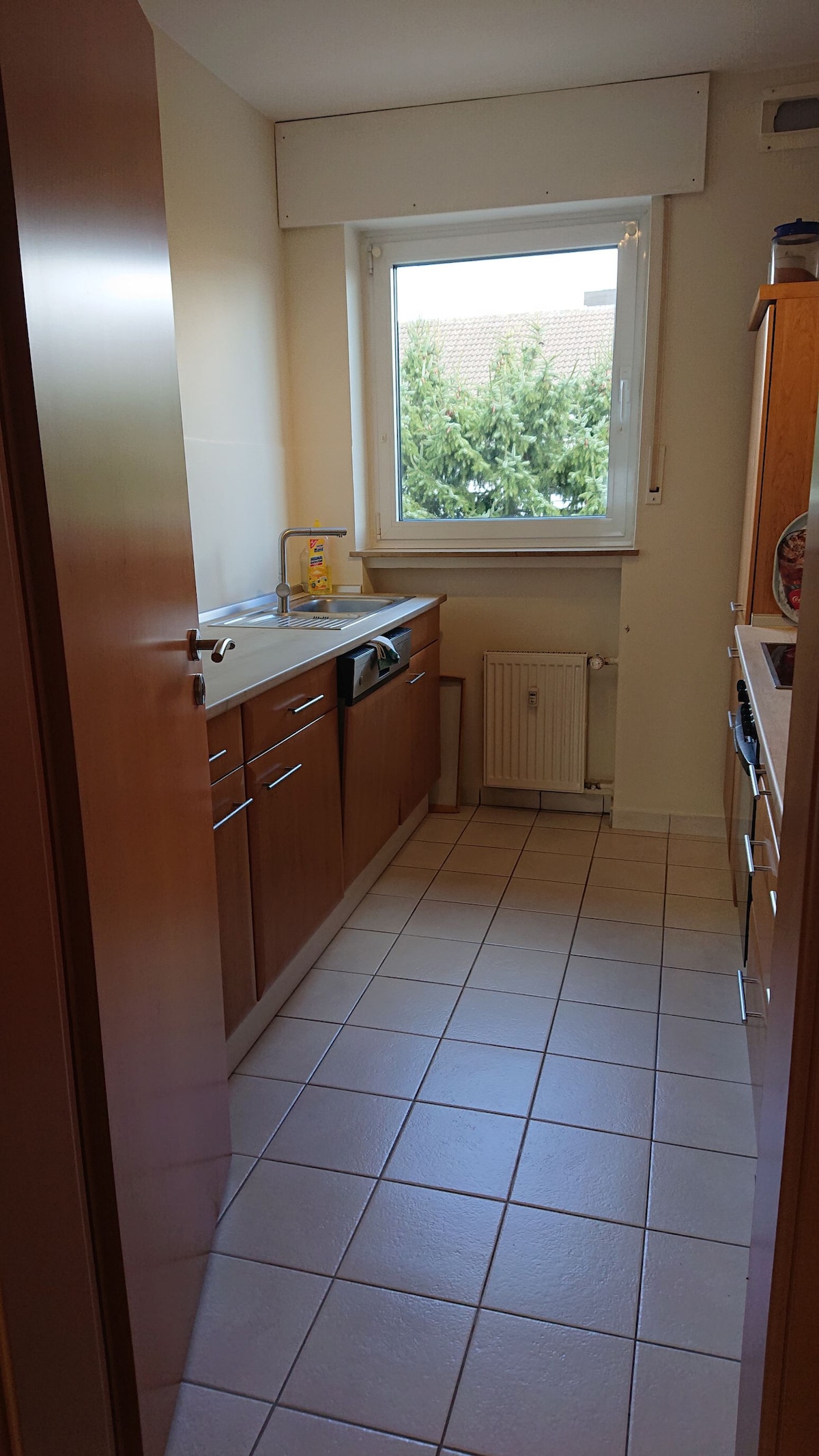 The kitchen features wooden cabinetry with a variety of storage options. A window allows natural light to illuminate the space, with a view of greenery outside. The tiled floor presents a clean and practical cooking area, enhancing functionality.