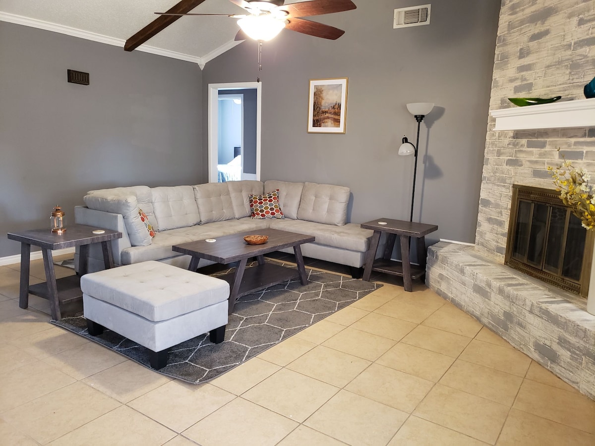 A spacious living area features a light gray sectional sofa with decorative pillows. Two dark wooden coffee tables are positioned in front, accompanied by a light gray ottoman. A stone fireplace adds a focal point, with a framed artwork displayed on the wall.