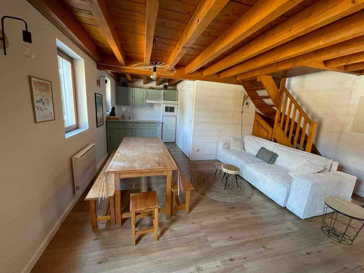 An inviting living space features exposed wooden beams and a light-toned wood floor. A spacious dining table is paired with several chairs, while a white sofa offers seating comfort. The kitchen is visible in the background, equipped with essential appliances, and a staircase leads to the upper level.