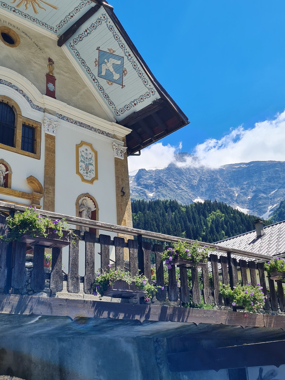 A charming building features ornate architectural details, with painted decorations and a wooden balcony adorned with colorful flowers. A majestic mountain backdrop is visible, framed by green trees under a clear blue sky, contributing to the serene setting.