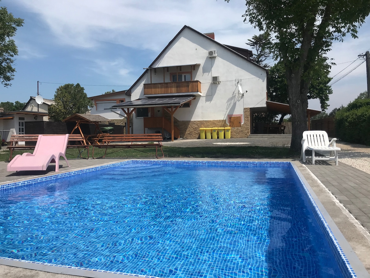 A swimming pool is set within a paved area, reflecting clear blue water under a bright sky. Wooden benches and a pink sun lounger are positioned nearby. The apartment building features a combination of wood and stucco construction, with a terrace extending from the upper level.