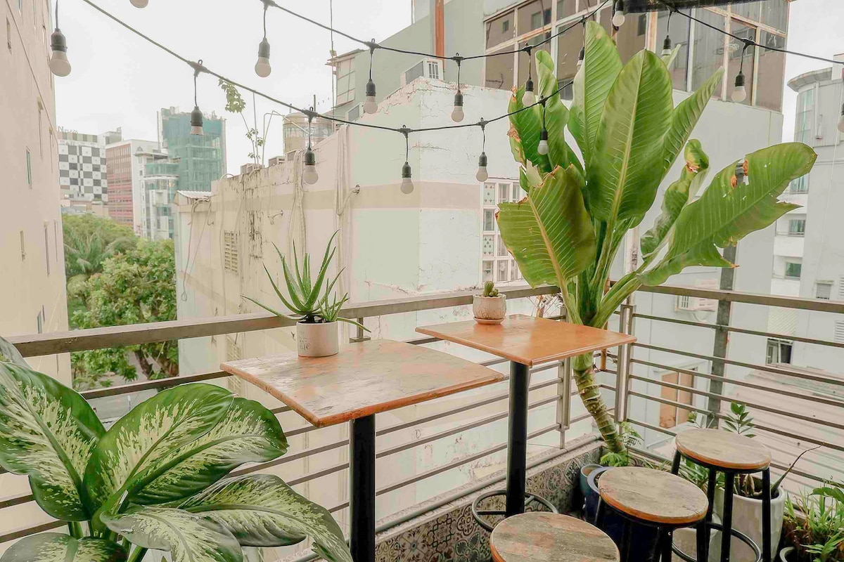 A balcony is seen with two wooden tables surrounded by potted plants. Strings of lights hang above, creating a cozy ambiance. The view includes buildings and greenery in the background, suggesting a lively urban environment.