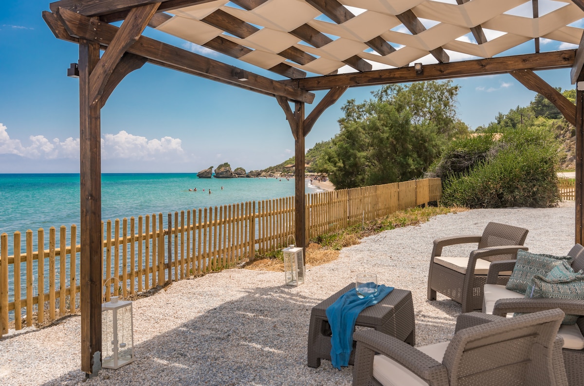 A shaded seating area with comfortable chairs and a table is positioned under a wooden pergola. The view of a soft sandy beach and clear blue waters of the Ionian Sea is visible in the background, framed by lush greenery along the shoreline.