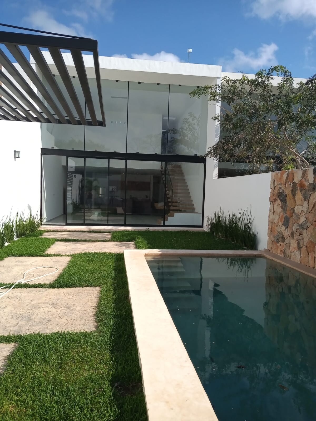 A modern two-story home is depicted, featuring large glass doors that provide a view of the interior. A stone pathway and verdant grass border a tranquil pool, reflecting the blue sky. Lush greenery frames the property, enhancing the serene outdoor space.