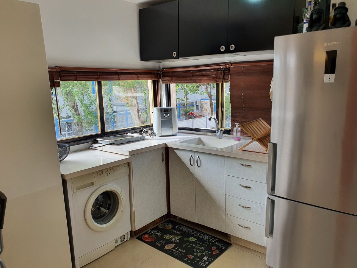 A bright kitchen space is presented with wood accents and large windows allowing natural light. A stainless steel refrigerator stands adjacent to a white countertop with a sink. A washing machine is situated to the left, and kitchen utensils are organized neatly nearby.