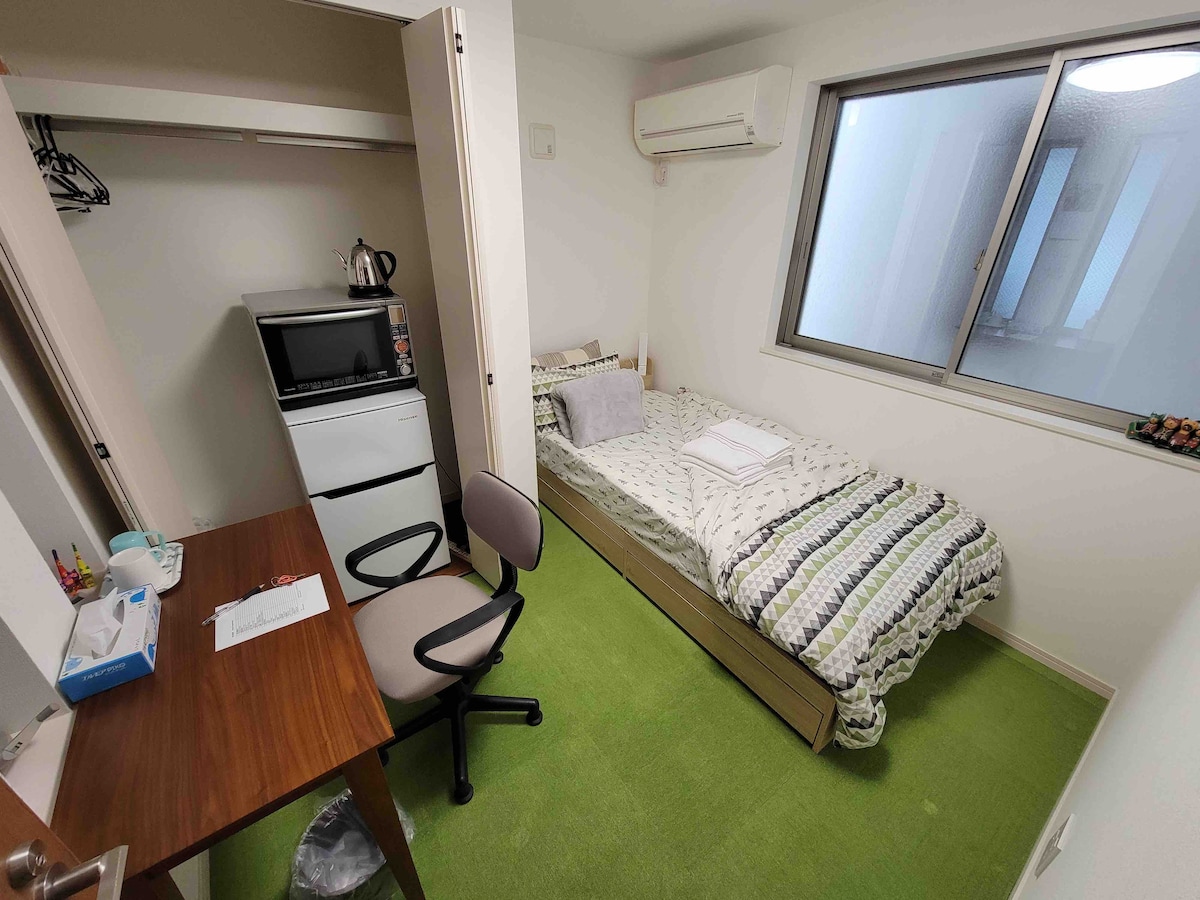 A compact bedroom features a single bed with linens neatly arranged. A desk with a chair is positioned beside a mini-fridge and a closet. Natural light enters through a window, providing a sense of openness to the green carpeted floor.