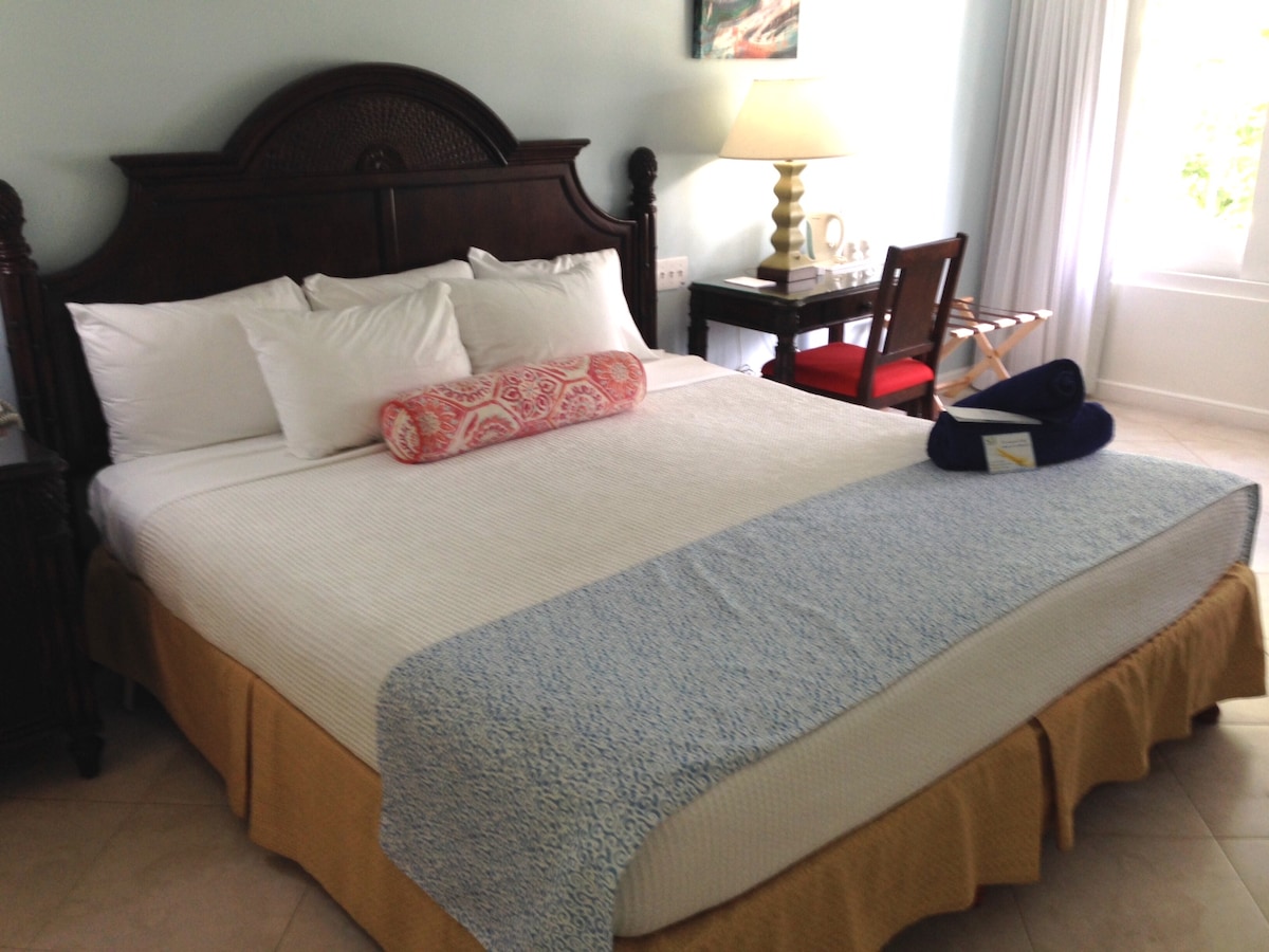 The spacious sleeping area features a queen bed with white linens and decorative pillows. A bedside lamp casts soft light, and a chair with a small table is positioned nearby. Natural light enters through a window with sheer curtains, enhancing the room's bright ambiance.