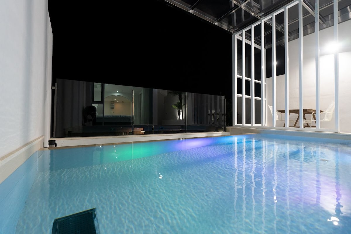 A private indoor swimming pool is visible, featuring calm waters illuminated by soft lighting. Floor-to-ceiling glass walls create a seamless connection to the surrounding space, while modern seating is positioned nearby, enhancing the area's open and airy feel.