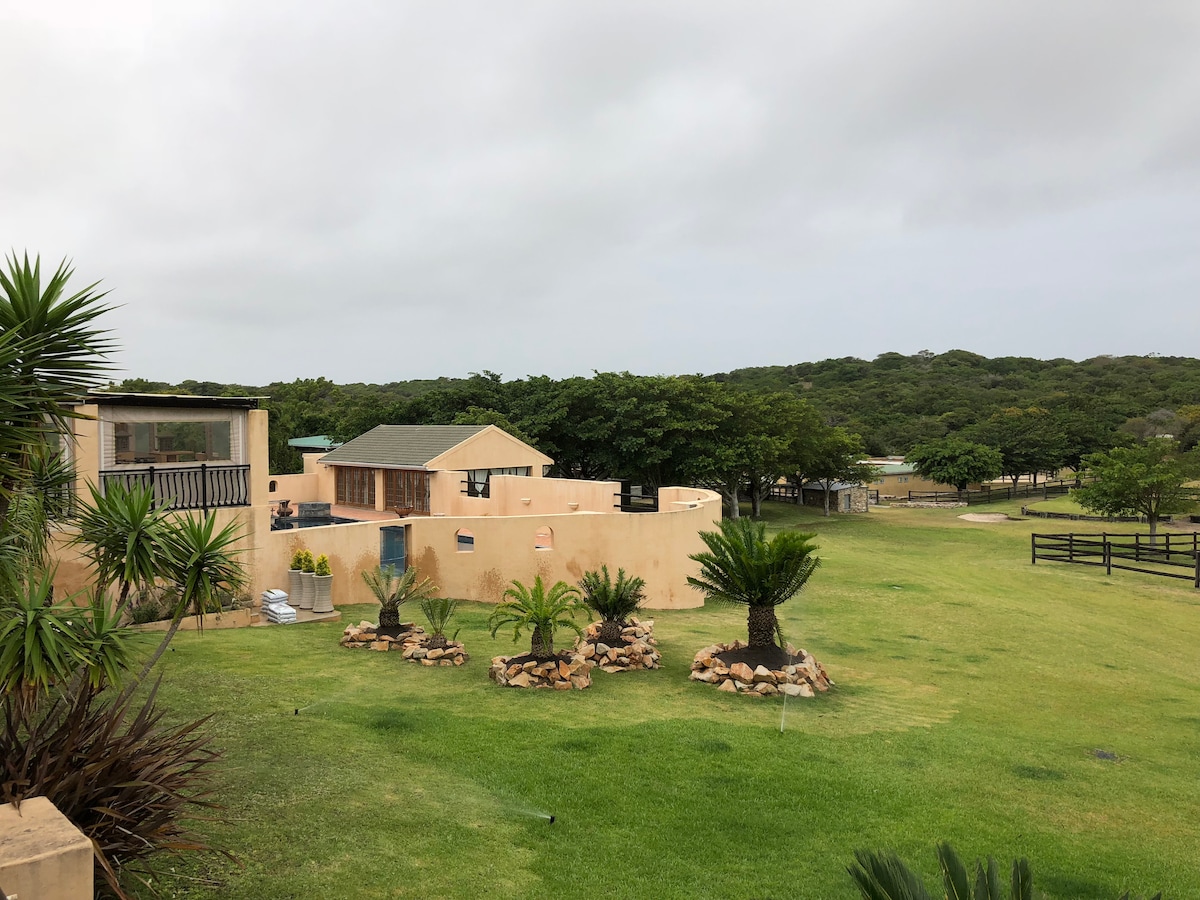 A spacious property is surrounded by lush green grass and palm trees, with a well-maintained garden area featuring decorative stones. The main building is complemented by various structures, set against a backdrop of trees that enhance the serene atmosphere.