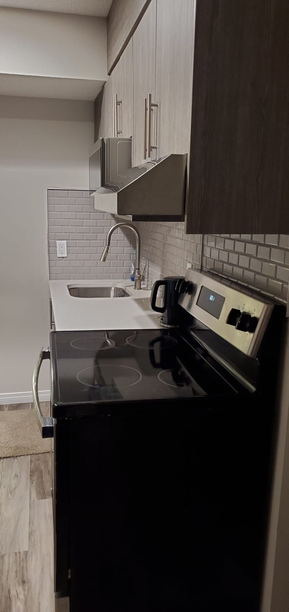 A compact kitchen area features modern cabinetry and a spacious counter. An electric stove is positioned beside a stainless-steel sink. Subtle tile backsplash enhances the design, while a kettle sits on the counter, ready for use. The overall aesthetic is clean and functional.