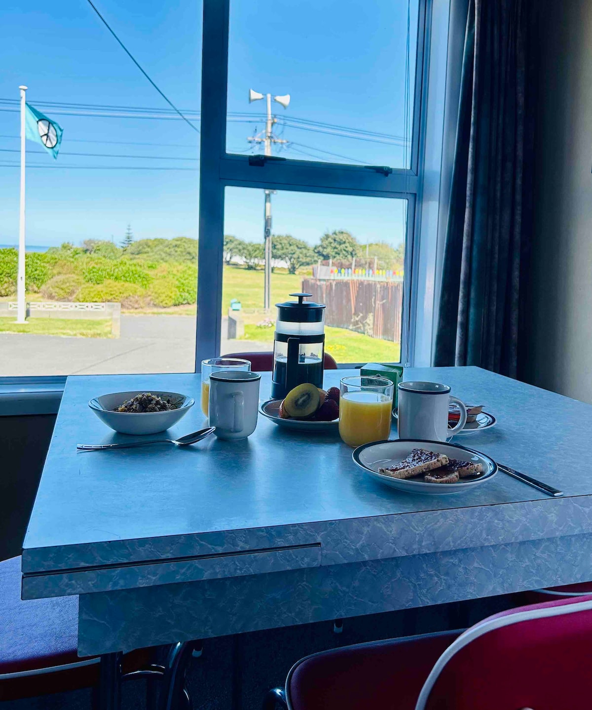 A retro-style dining table is set with bowls of cereal, a cup, a glass of juice, and a French press. The view through the window includes green fields and a flag. Bright light illuminates the scene, enhancing the inviting breakfast setting.