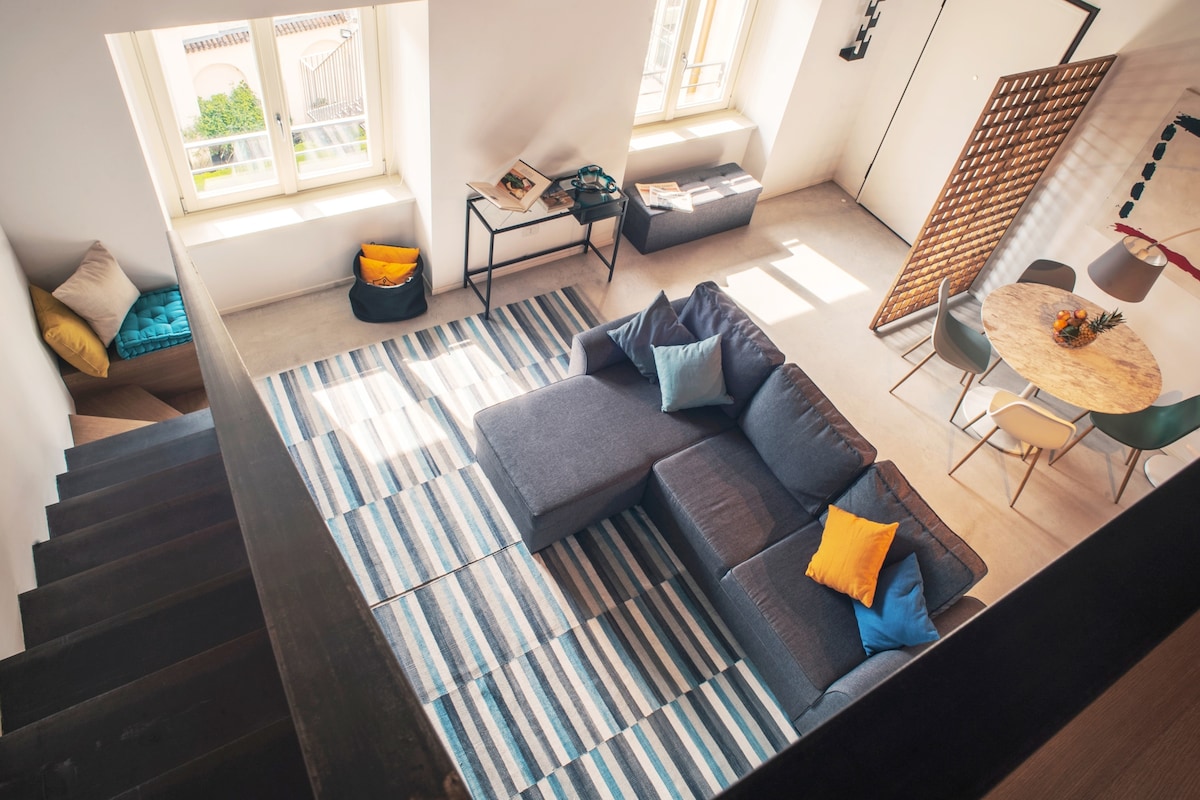 A spacious living area is visible from above, featuring a large sectional sofa with bright cushions. A circular dining table is set with colorful chairs. A striped area rug adds texture, while sunlight filters through the large windows, enhancing the openness of the space.