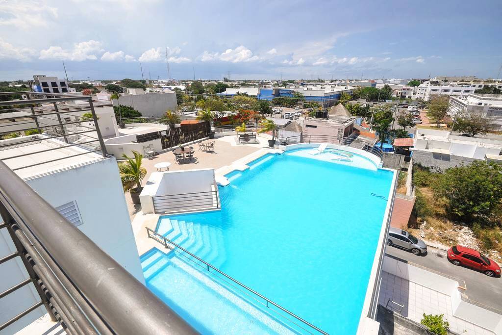 The rooftop pool area is large and inviting, featuring a spacious pool with steps leading in from the surrounding sundeck. Lounge chairs are positioned around the pool, offering an ideal space for relaxation. A panoramic view of Playa del Carmen’s skyline is visible in the background.