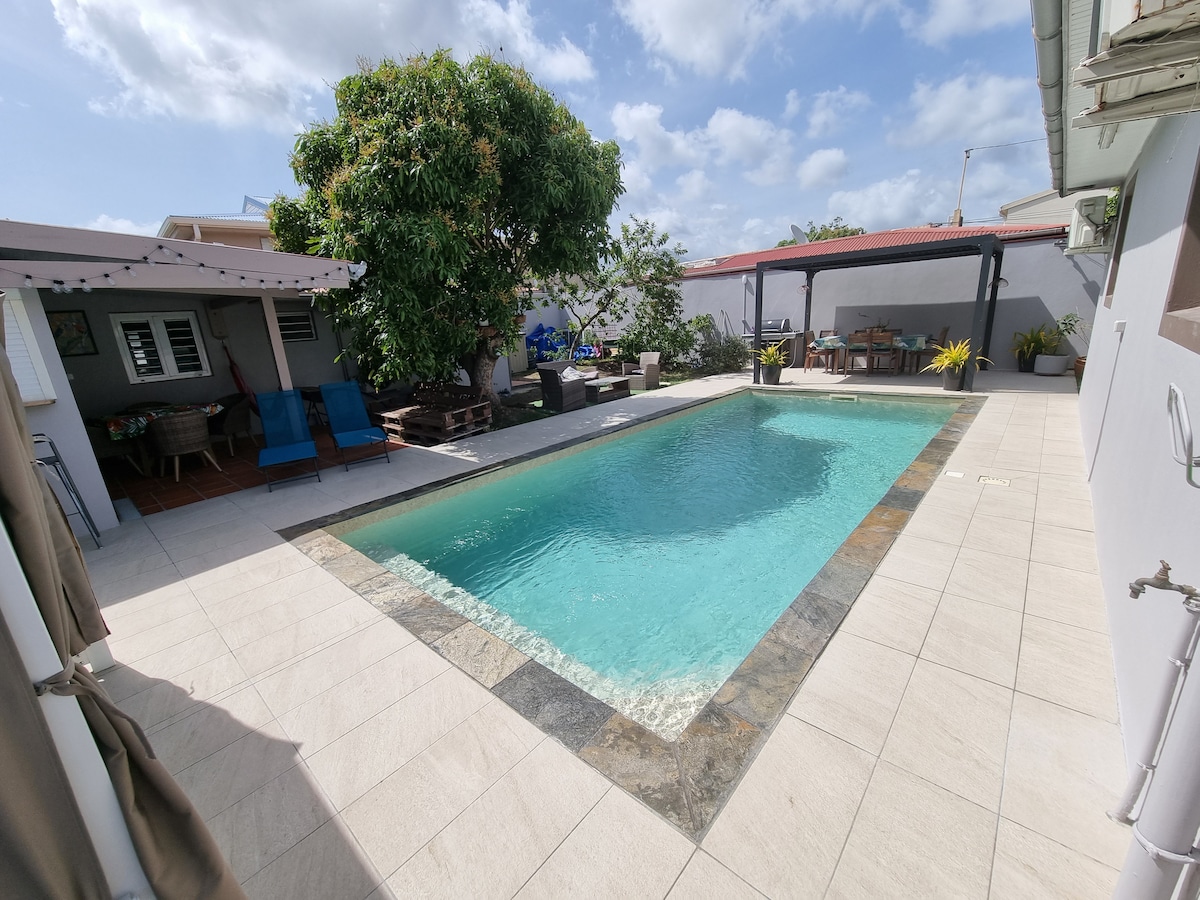 The outdoor area features a spacious swimming pool surrounded by a stone border. Lounge chairs are positioned nearby, and a shaded seating area offers a place to relax. Colorful plants enhance the inviting atmosphere, while a large tree provides natural shade.