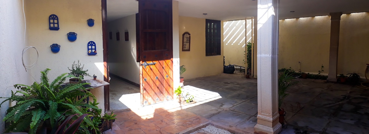 An entryway features a blend of stone and tiled flooring, illuminated by natural light. Lush greenery decorates the sides, with potted plants adding a touch of nature. Wooden doors lead to an inner area, enhancing the welcoming atmosphere of the space.