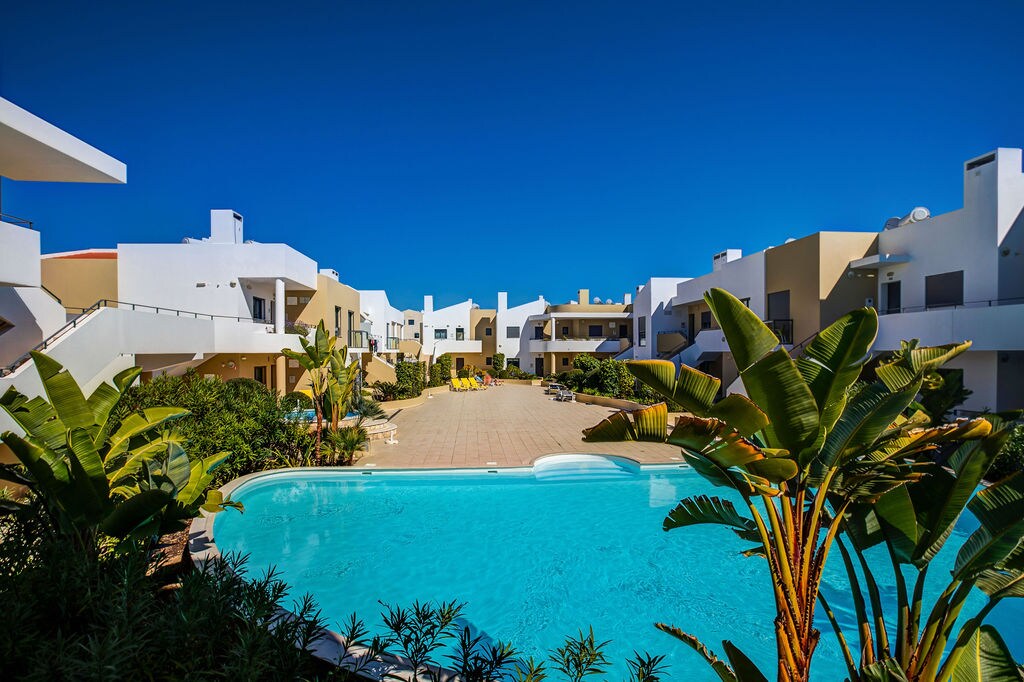 The image displays a serene swimming pool surrounded by lush greenery and modern apartment buildings. A clear blue sky sets the backdrop, while sun loungers and umbrellas are positioned along the poolside, enhancing the inviting atmosphere of the complex.