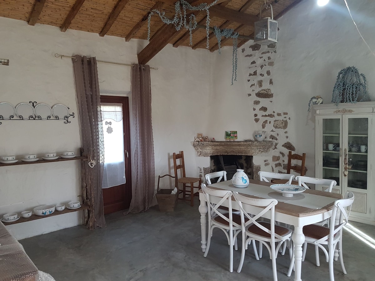 A cozy dining area is featured with a wooden table surrounded by white chairs. The stone fireplace adds warmth, while shelves hold decorative bowls. Natural light enters through a door draped with sheer curtains, and rustic elements create a charming ambiance.
