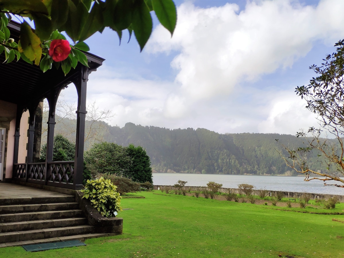The image captures a scenic view from a porch, showcasing a lush green lawn leading to a tranquil lake. Mountains rise in the background under a partly cloudy sky. A flowering plant with vibrant leaves is visible in the foreground, enhancing the natural setting.