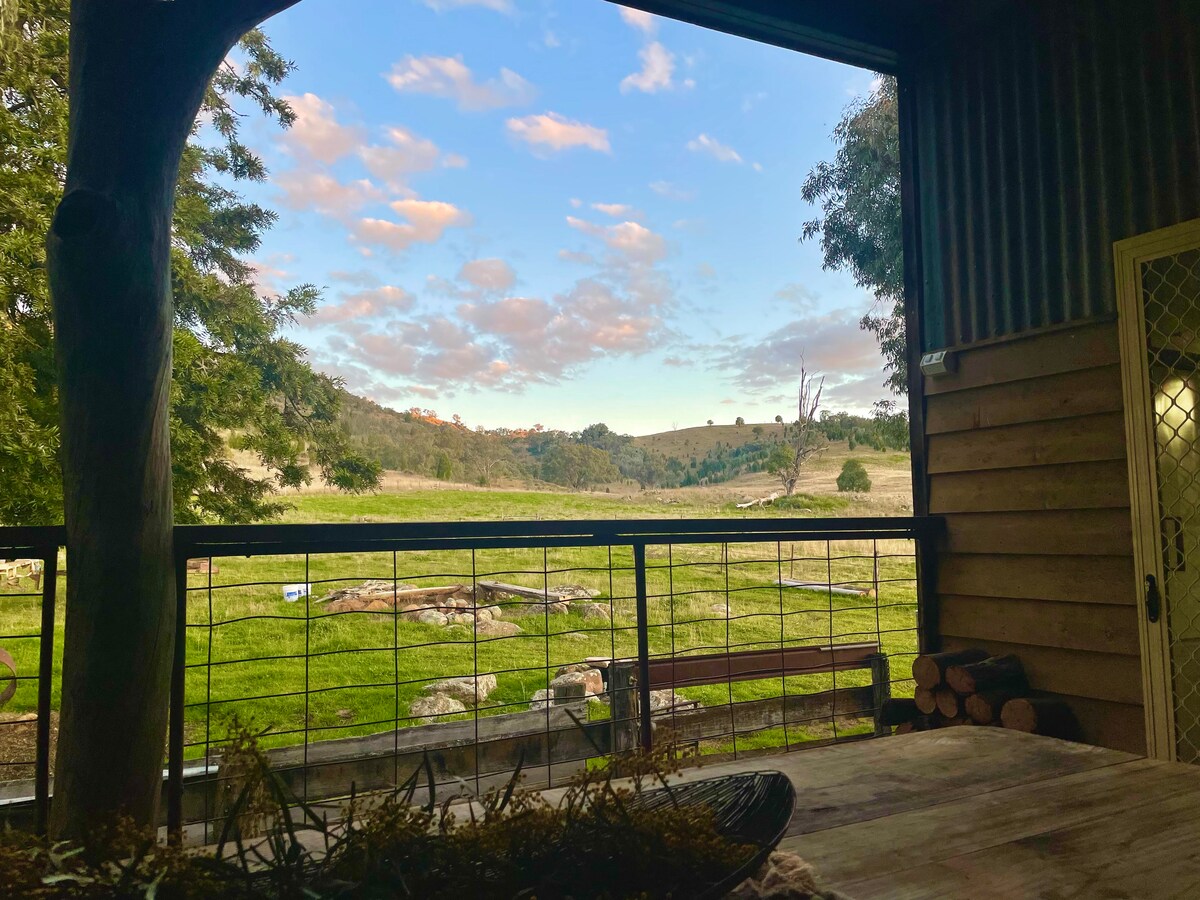 A serene outdoor view is captured from the porch, showcasing rolling hills and a vibrant sky filled with soft clouds. A wooden table with natural accents is visible in the foreground, while a fire pit area is seen in the distance, inviting relaxation.