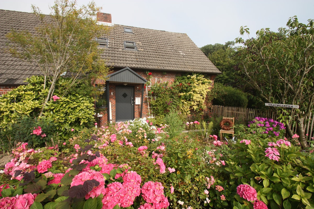 The exterior of the house is complemented by a vibrant garden filled with colorful flowers, particularly pink hydrangeas and lush greenery. A walkway leads to the entrance, framed by blooming plants and shrubs, enhancing the welcoming appearance of the property.