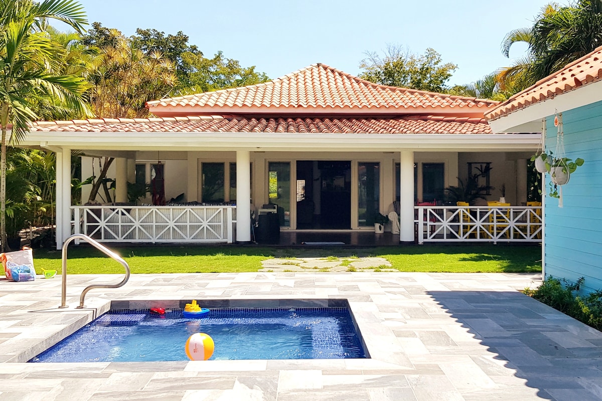 A charming villa is set amidst lush greenery, featuring a covered terrace with plentiful seating. A small, inviting hot tub is visible in the foreground, surrounded by light-colored stone tiles. A playful beach ball floats on the water, enhancing the serene outdoor space.