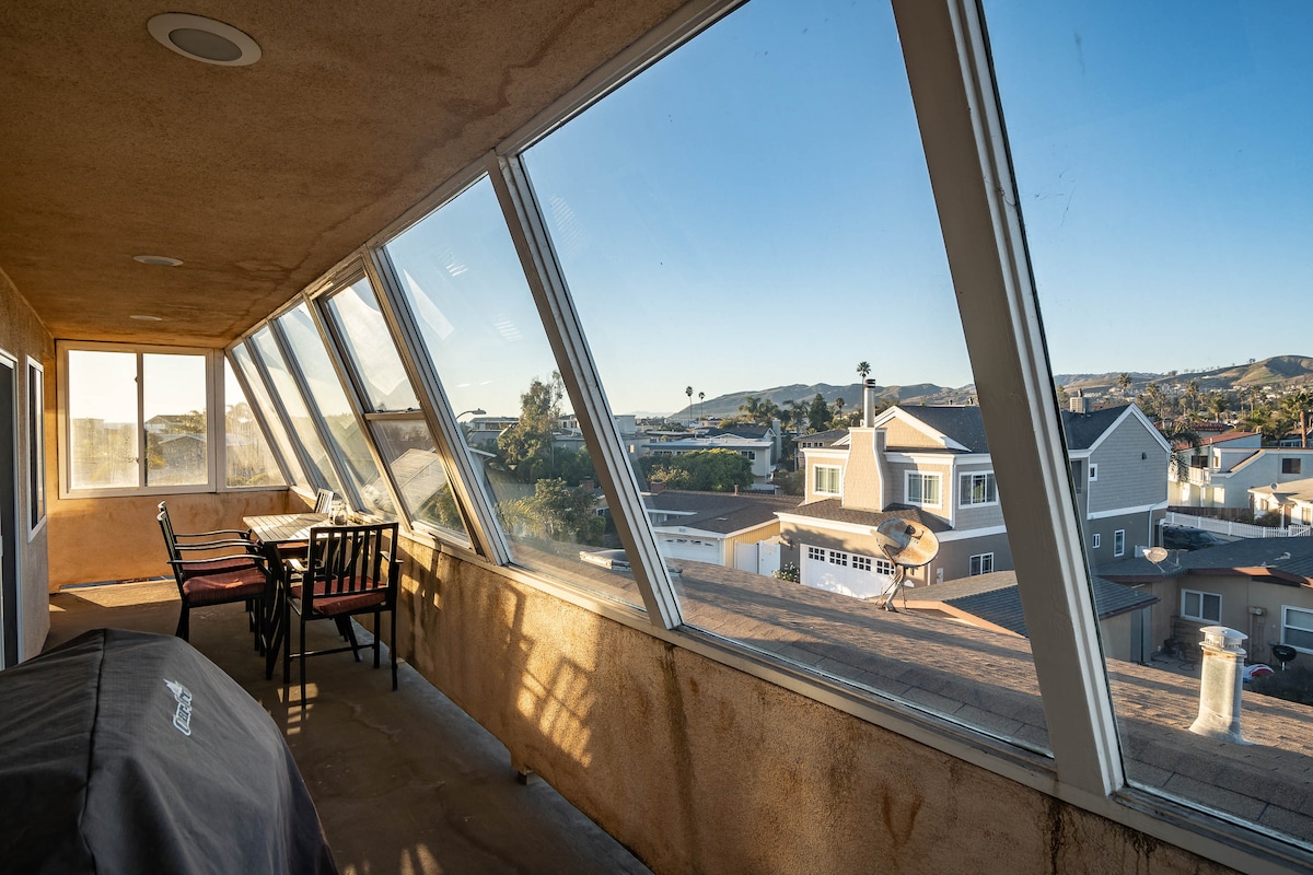 A sunlit enclosed patio features large angled windows offering panoramic views of the surrounding area. A dining table with chairs is visible, alongside a barbecue grill, creating an inviting space for relaxation and gatherings.