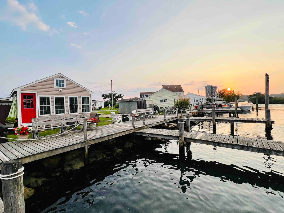 A waterfront cottage is positioned alongside a wooden dock, with a vibrant red door contrasting the neutral façade. Octagonal benches and planters with flowers are visible on the grassy area. The scene captures a serene sunset over calm waters, enhancing the peaceful environment.