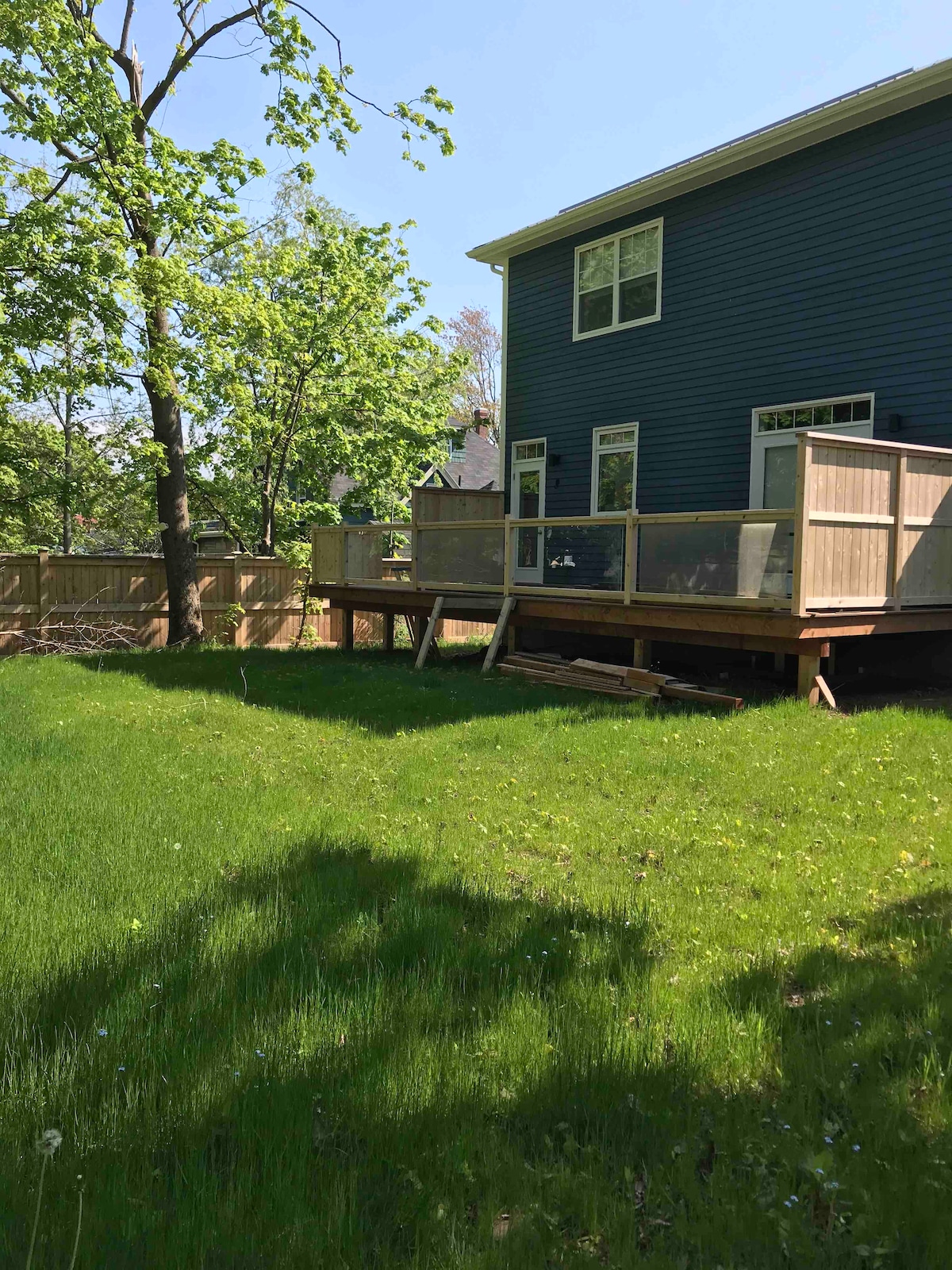 A spacious wooden deck is elevated above a lush green lawn, surrounded by trees and a wooden fence. The blue exterior of the house contrasts with the vibrant greenery, creating a serene outdoor space for relaxation.