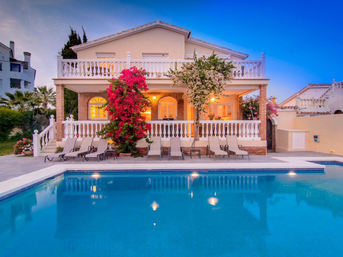 The villa is showcased from the poolside, featuring a pastel facade with a covered terrace adorned with flowering plants. Sun loungers are arranged around the private pool, reflecting the blue sky and providing a serene view of the surrounding garden.