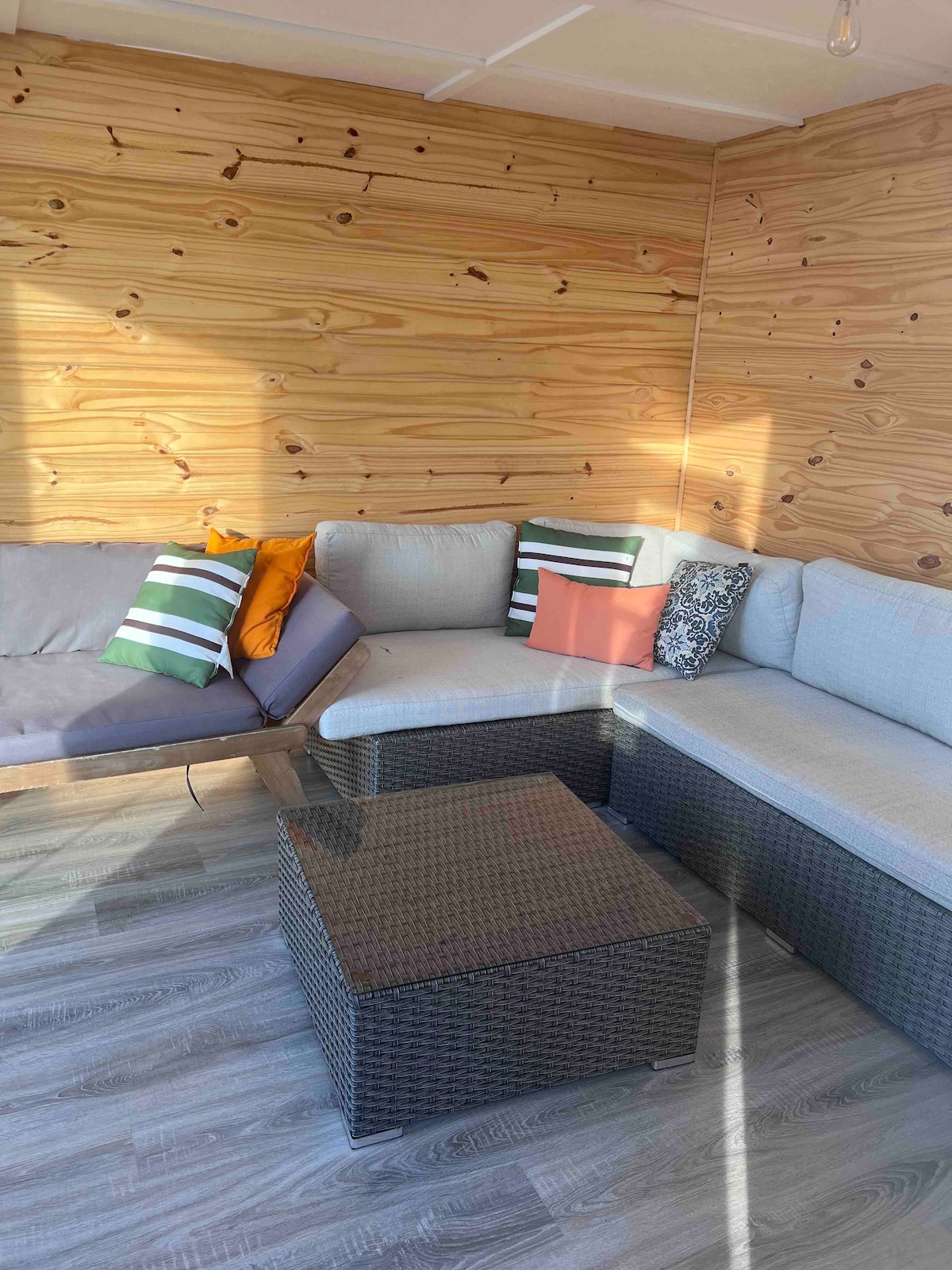 A cozy seating area features two grey sectional sofas adorned with colorful cushions in green, orange, and patterned designs. A glass-top coffee table sits in the center, surrounded by light wood paneling that enhances the natural ambiance. Sunlight illuminates the space.