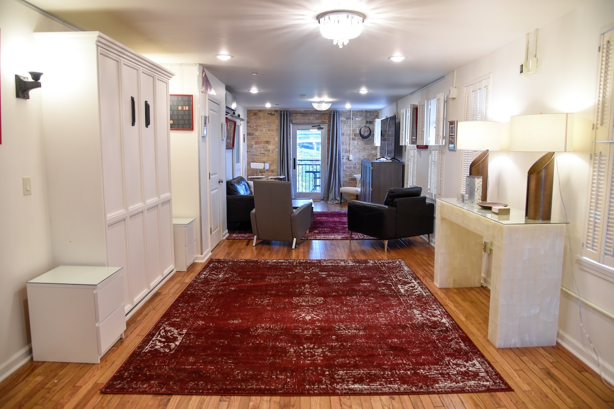 An open studio space features hardwood flooring and a large area rug in deep red tones. Seating areas consist of modern chairs and a combination of cabinetry providing storage. Natural light is welcomed through multiple windows, and a balcony view is visible at the far end.