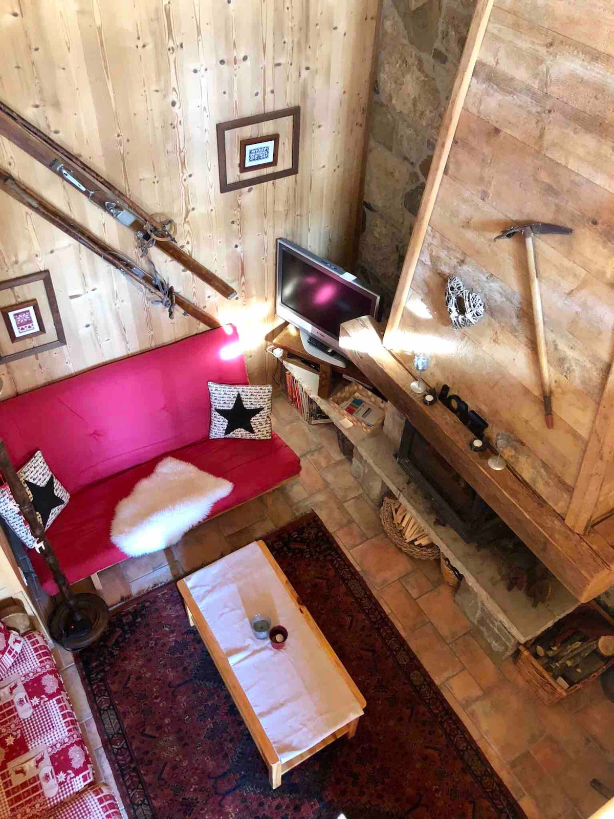 An inviting living area with wooden walls showcases a bright red sofa adorned with decorative cushions. A rustic wooden coffee table sits centrally, with a patterned area rug beneath. A television, ski equipment, and decorative items are arranged on a shelf above the cozy fireplace.