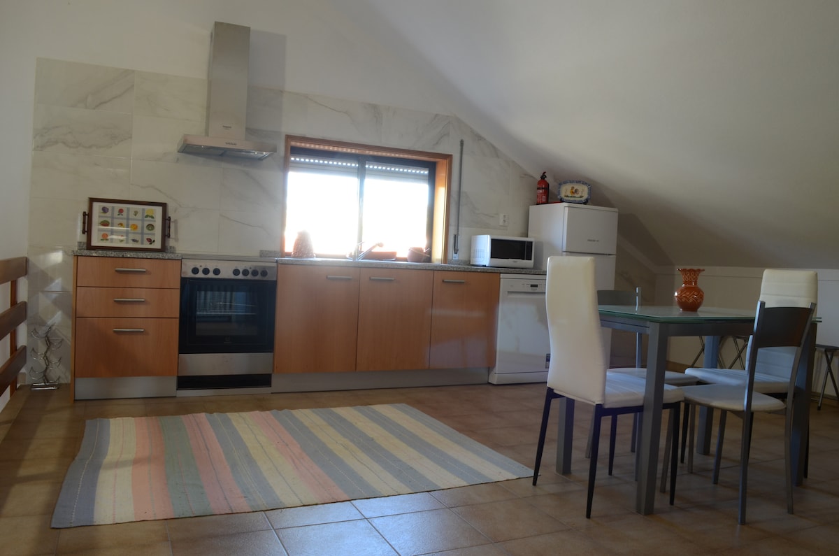 A modern kitchen features wooden cabinetry and a stainless steel oven. A dining table is set with four chairs, accompanied by a colorful striped rug on the tiled floor. Natural light filters through a window, bringing warmth to the space.
