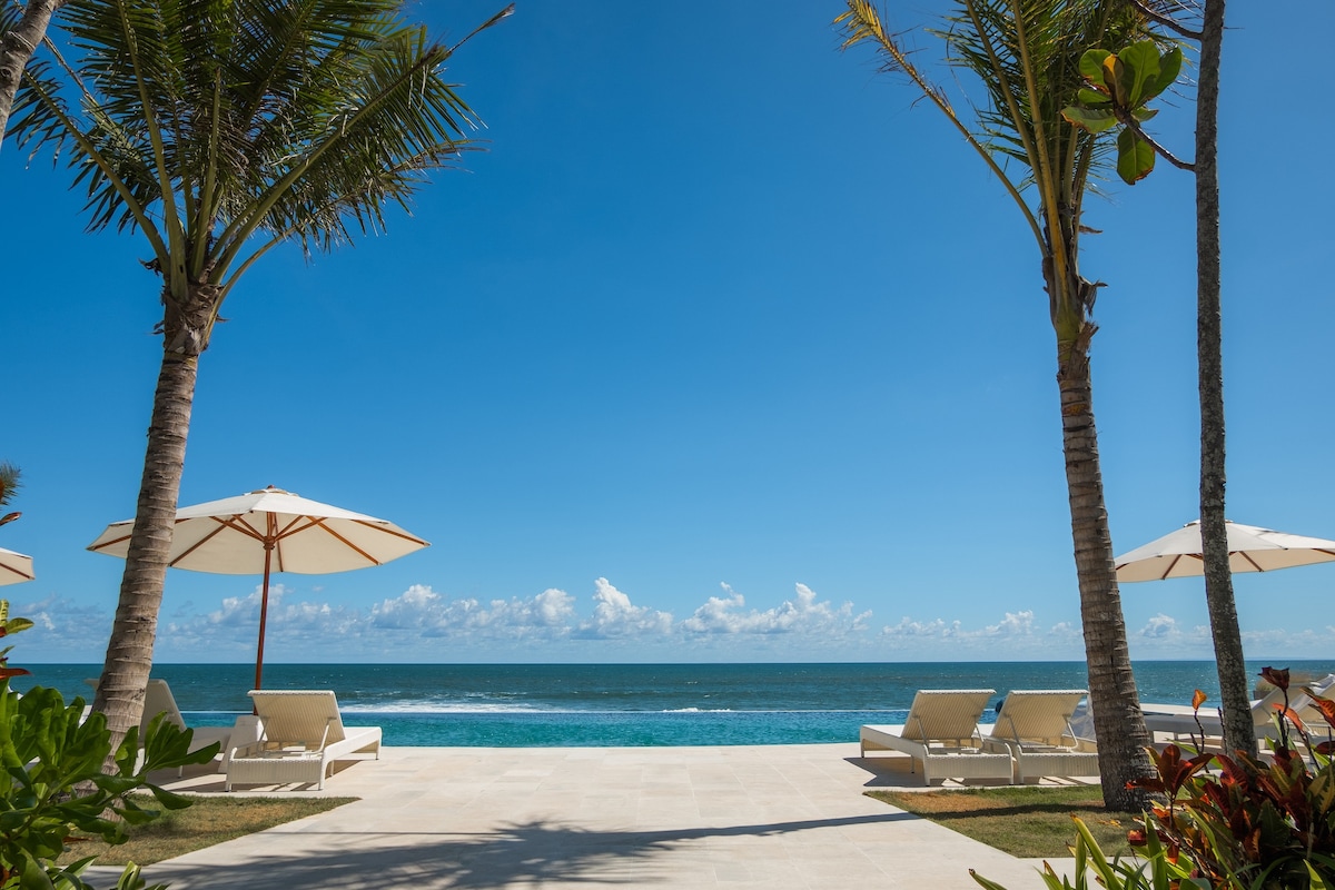 A view of the ocean is framed by two palm trees, standing beside lounge chairs beneath large umbrellas. The ocean water reflects the clear blue sky, while a smooth walkway extends toward the beach, creating an inviting outdoor space.