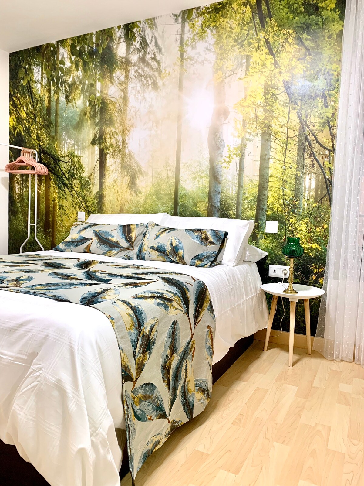 The bedroom features a large bed adorned with a decorative cover showcasing a nature-inspired pattern. A vibrant mural depicting a sunlit forest creates a serene backdrop. A small nightstand is visible beside the bed, complemented by a clothing rack in the corner.