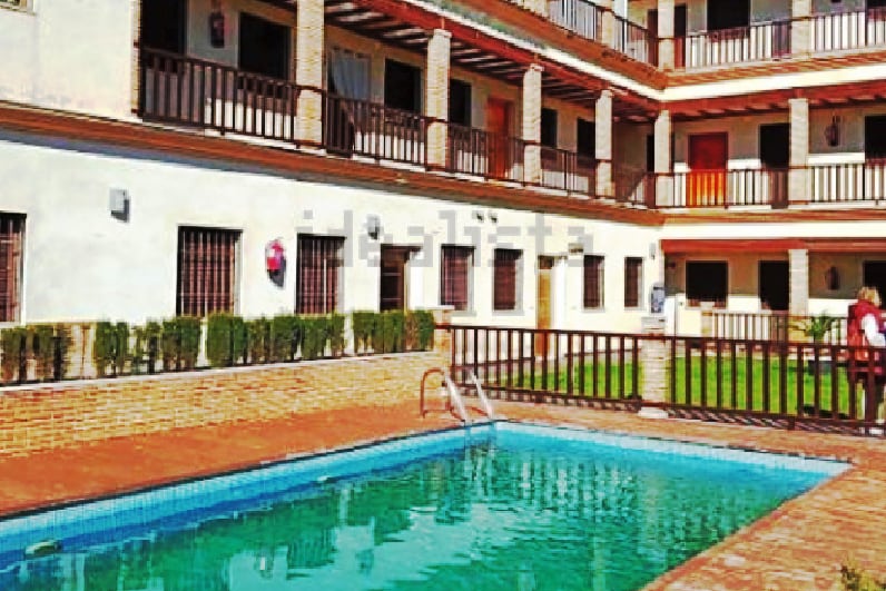 A rectangular outdoor pool is surrounded by a tiled patio and bordered by well-maintained greenery. The building features multiple balconies with wooden railings, creating a layered visual element against the sunny backdrop.