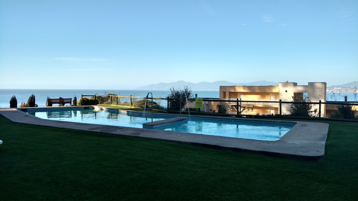 Two swimming pools are visible, set within well-maintained grass and surrounded by several landscaped areas. The ocean can be seen in the background, with distant hills enhancing the view. A lounge area with seating is present nearby, inviting relaxation.