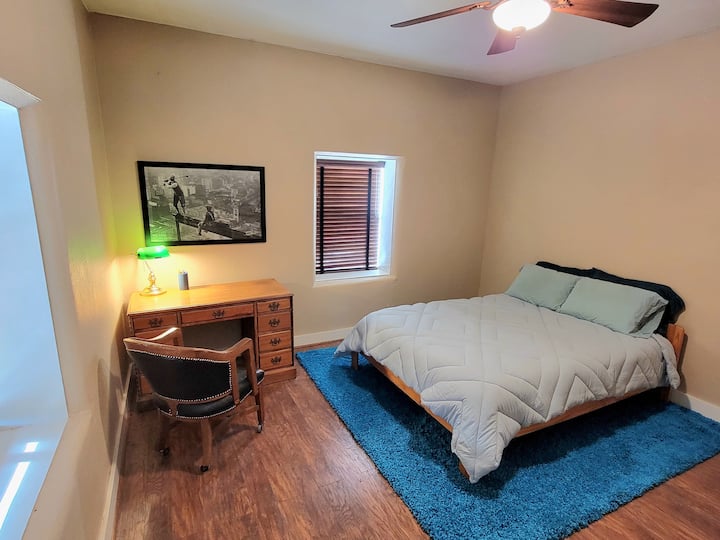 Dormitorio con cama tamaño queen, espacio de trabajo para escritorio y una pequeña biblioteca de libros con historia, historias y arte de Nuevo México.  ¡Y una lujosa alfombra turquesa!