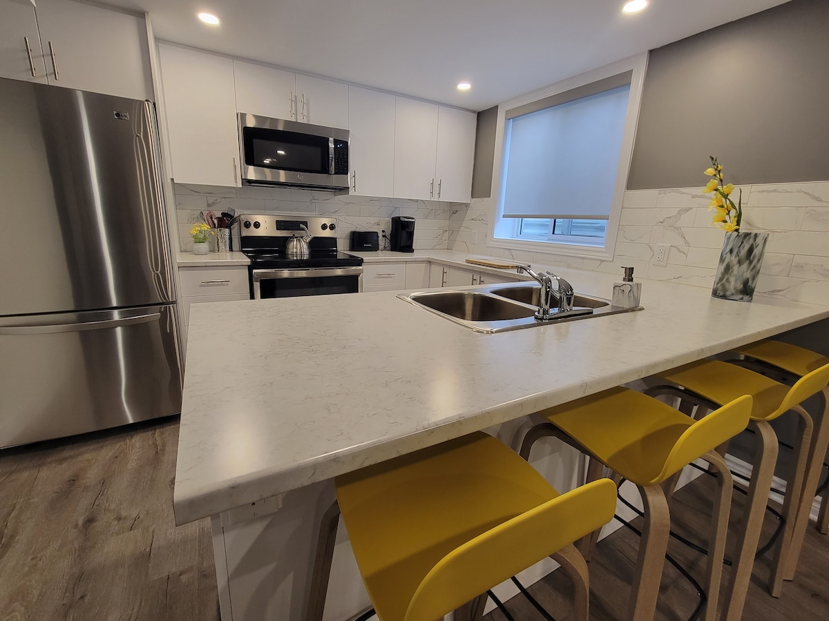 A modern kitchen features sleek white cabinetry and stylish stainless steel appliances. A spacious island with a light-colored countertop is equipped with three yellow bar stools. Natural light enters through a nearby window, enhancing the contemporary design.