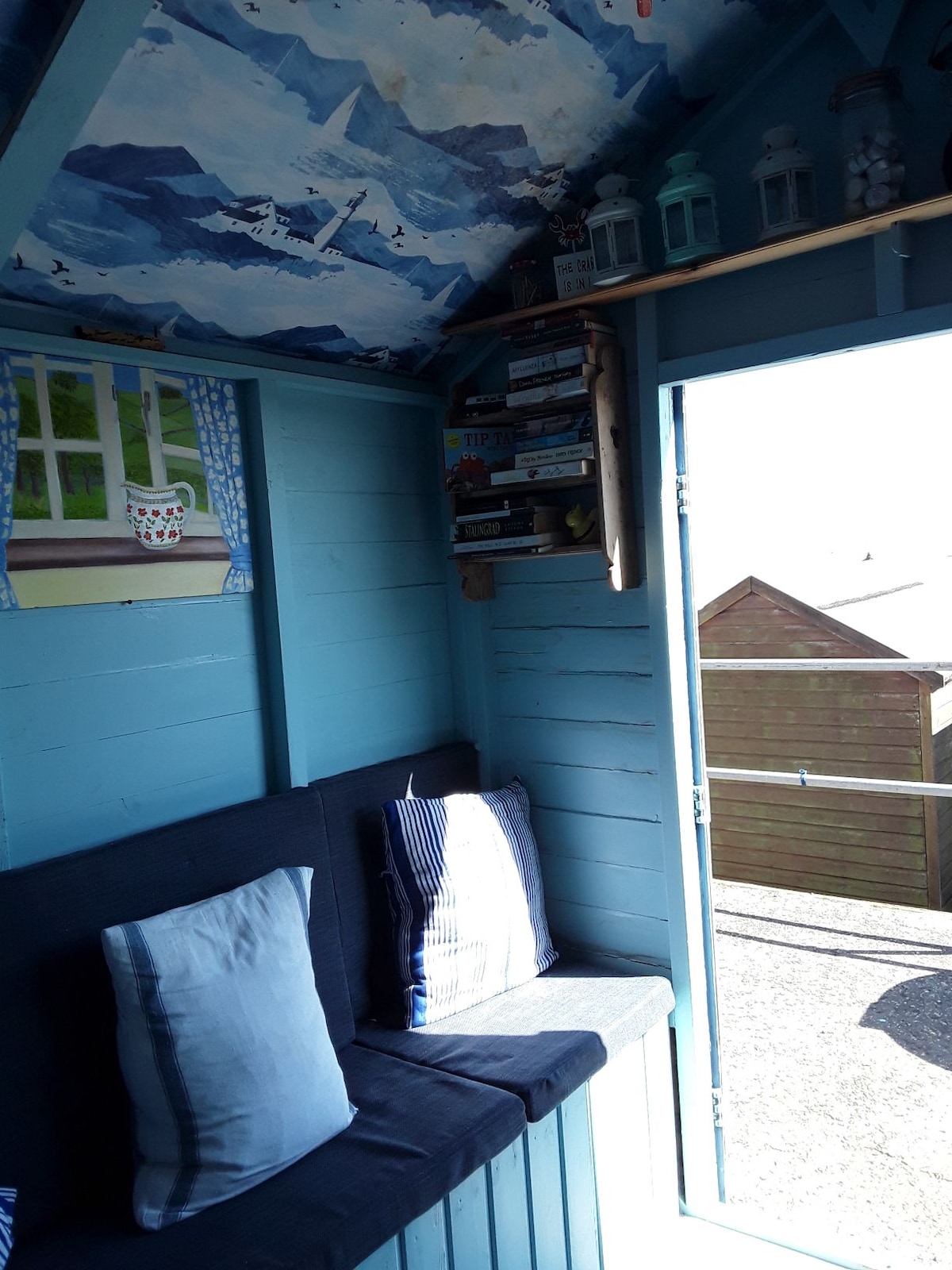 The interior of the beach hut shows a cozy seating area with built-in benches covered in striped cushions. A colorful mural featuring mountains is painted on the ceiling, and a shelf filled with books can be seen in the corner. Natural light enters through an open door, illuminating the space.